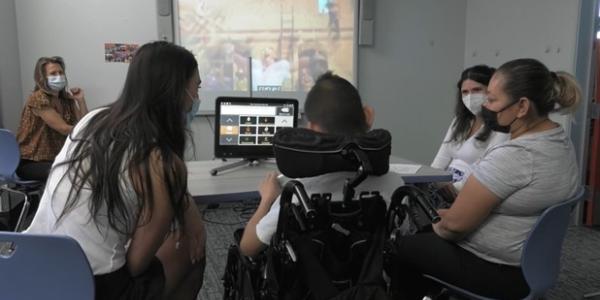 An adolescent in a wheelchair using eye gaze to access his AAC system, while his mother and two graduate clinicians show him comments on the AAC system about the movie Encanto  that’s playing on the screen. 