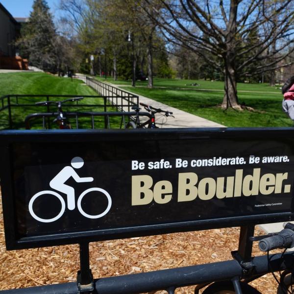 Student riding a bike past Be Boulder. sign