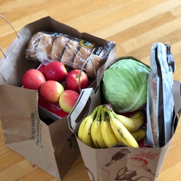 An overhead view of two paper bags sitting on a wood counter filled with grocery staples like produce and bread