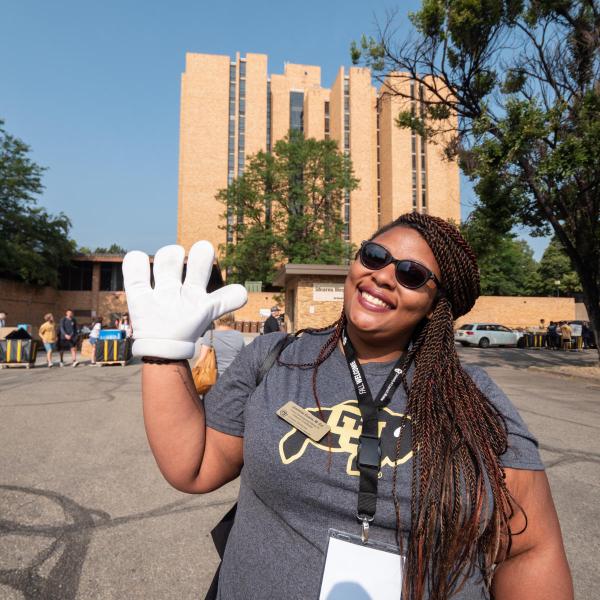 A female staff member wearing a CU Athletics shirt waves stands outside Will Vill waving at the camera with a cartoon hand