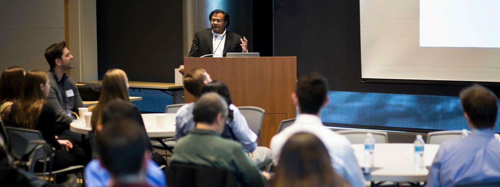 A man stands at a podium, enthusiastically speaking to a large group of students.