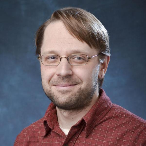 A portrait of Dr. David Bortz smiling. He has brown short hair, is wearing thin wire glasses, and is wearing a red button up shirt. He is sitting in front of a blue background.