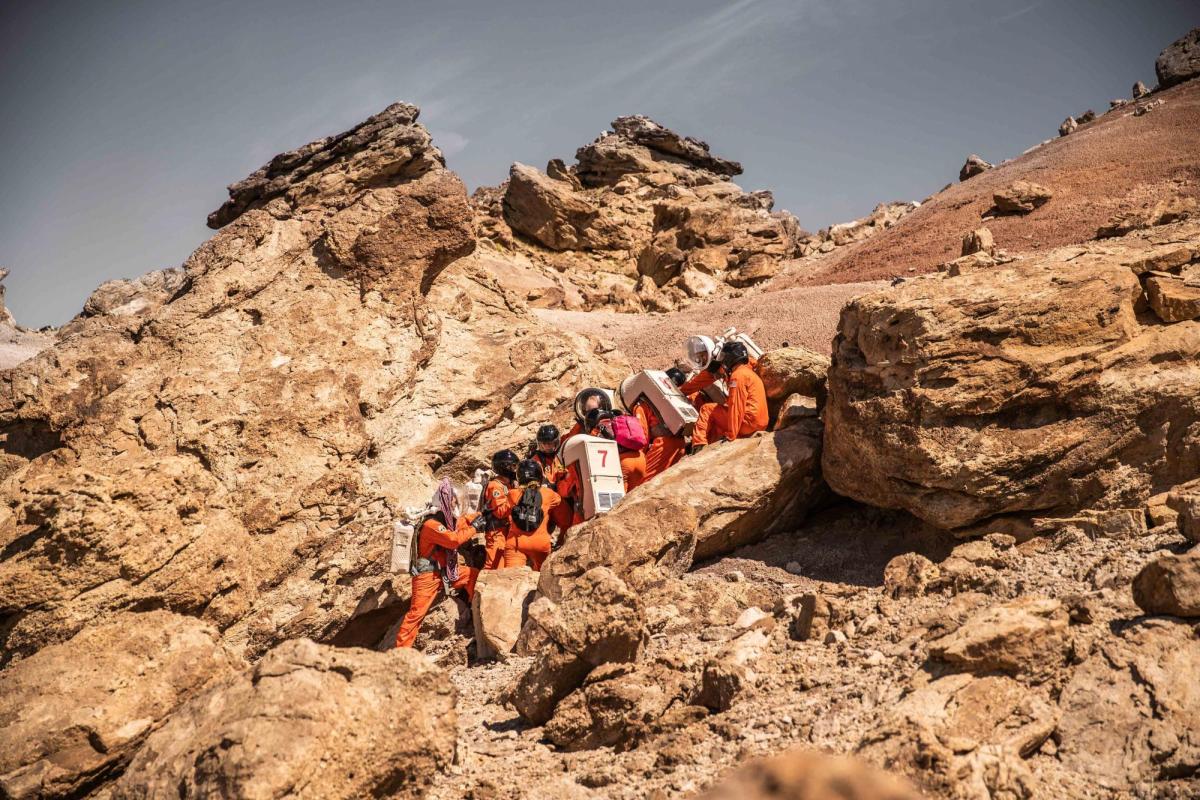 Group of people in orange space suits climbing hill in the desert