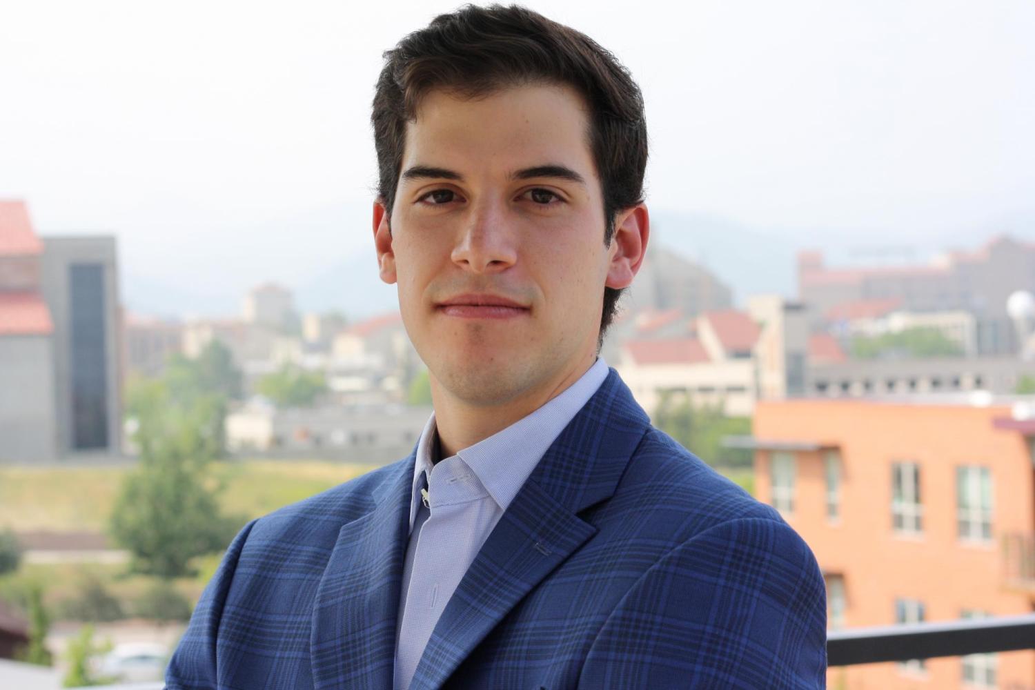 Adrian Gutierrez standing with arms folded in a blue suit. The background: Buildings in the distance with orange roofs.