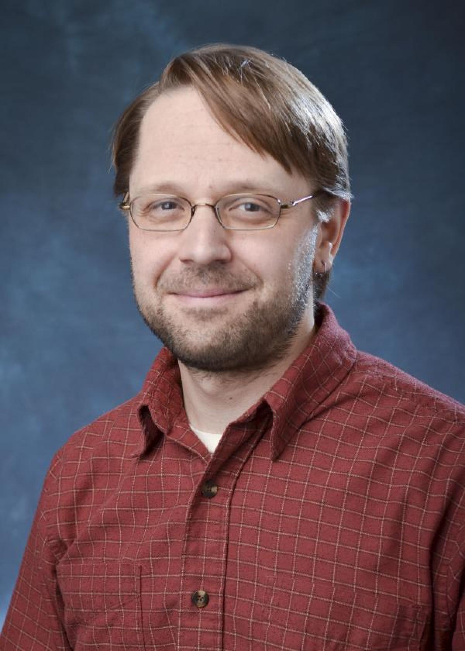 A portrait of Dr. David Bortz smiling. He has brown short hair, is wearing thin wire glasses, and is wearing a red button up shirt. He is sitting in front of a blue background.