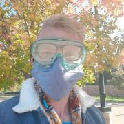 Assistant Professor Cresten Mansfeldt is standing outside in the sun. He is wearing eyeglasses that around surrounded by green protective goggles. He is also wearing a large grey mask that is covering his nose, mouth, and lower face. 