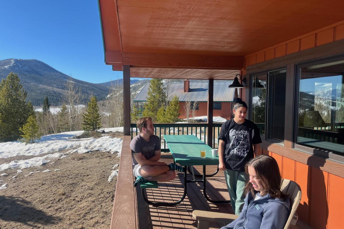 three people sitting on a cabin porch with a mountain in the background