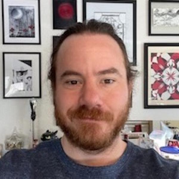 Selfie of a white male-presenting individual with long reddish-brown hair secured behind his hair and a beard.
