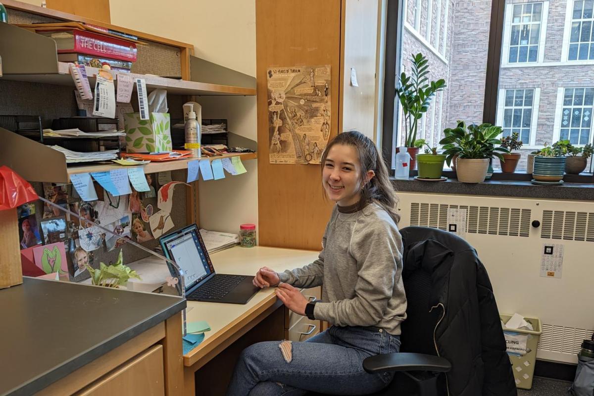 Light-skinned undergraduate student smiles and poses for a picture of her at her desk in a research lab.