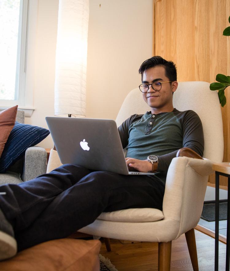 Male student in green shirt and jeans lounging in armchair with legs propped up, working on laptop