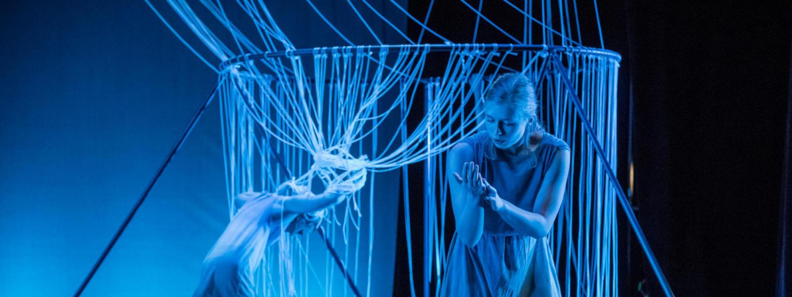 A dancer moves through a structure made of string while another looks at their hands