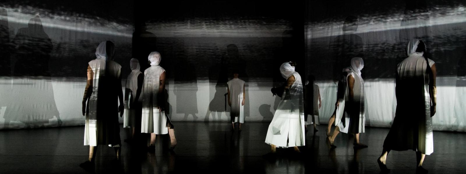 Several dancers walking on stage. Each wears a face covering. The backdrop is white and black stripes