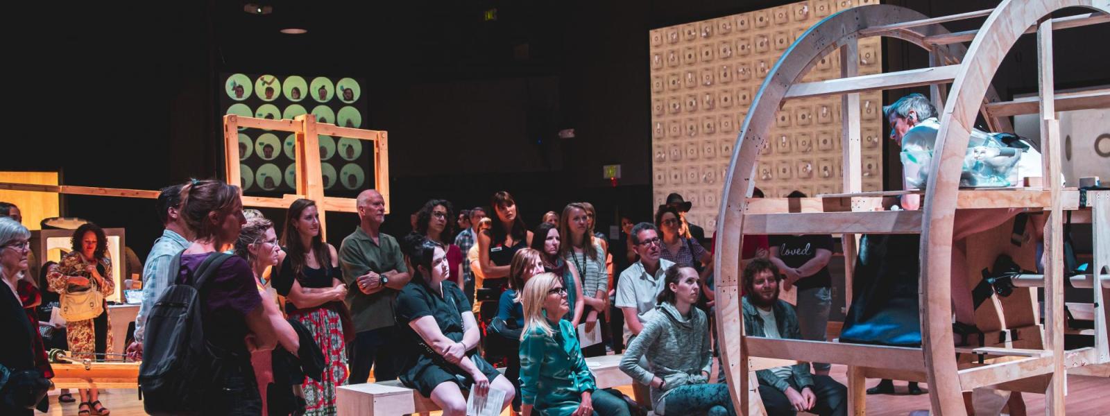 A performer speaks to an audience from on top of a large wooden wheel structure