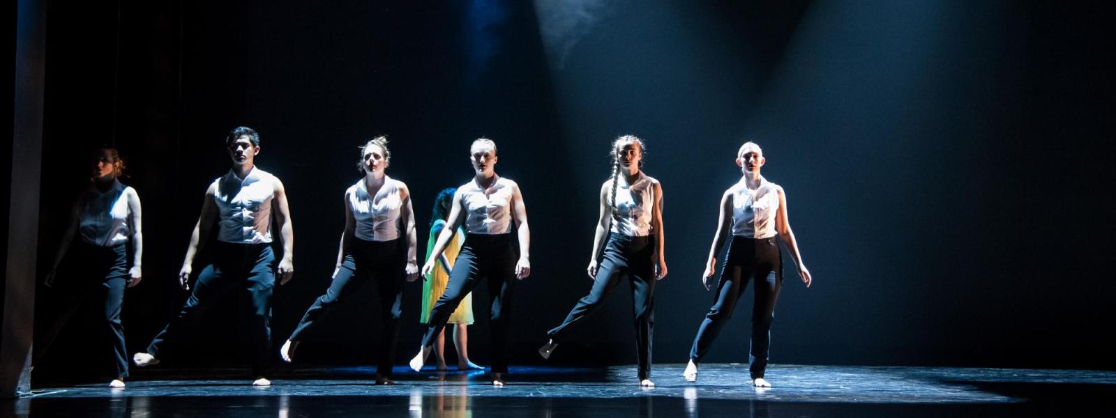 Six dancers stand in a row downstage in hazy light