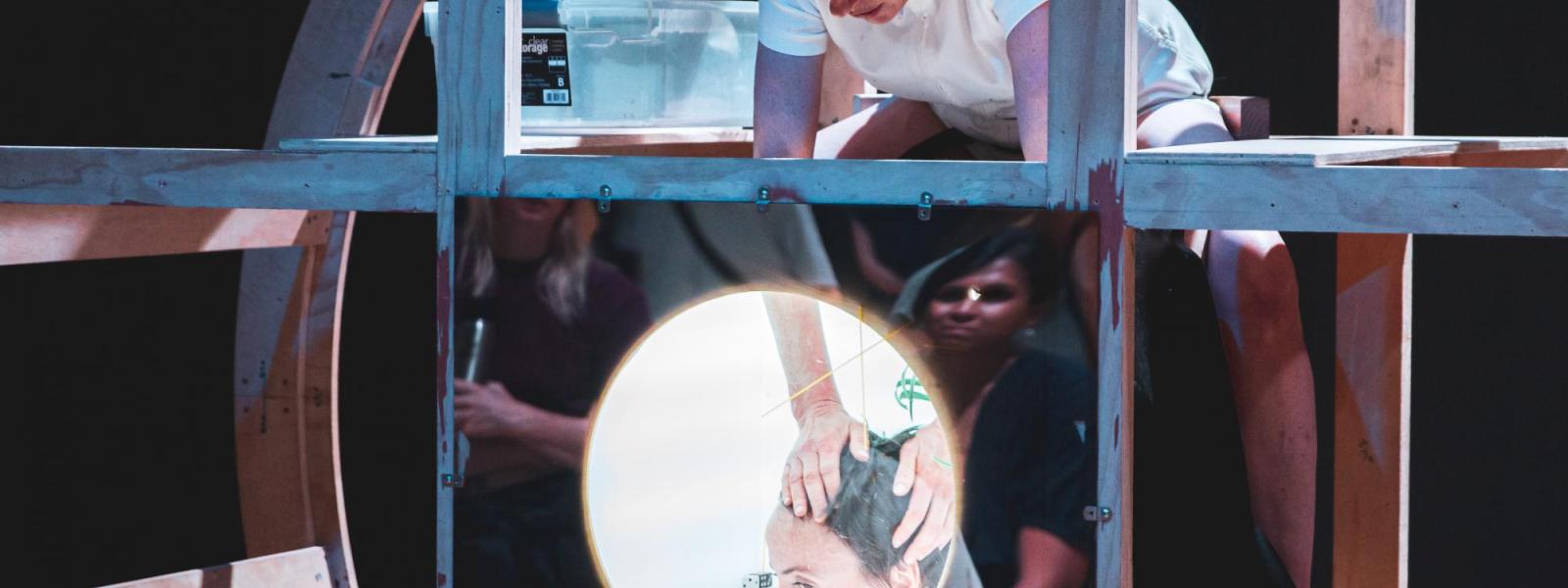 A performer puts her hands into a mirrored box to touch another performer's hair. Both are inside a huge wooden wheel.
