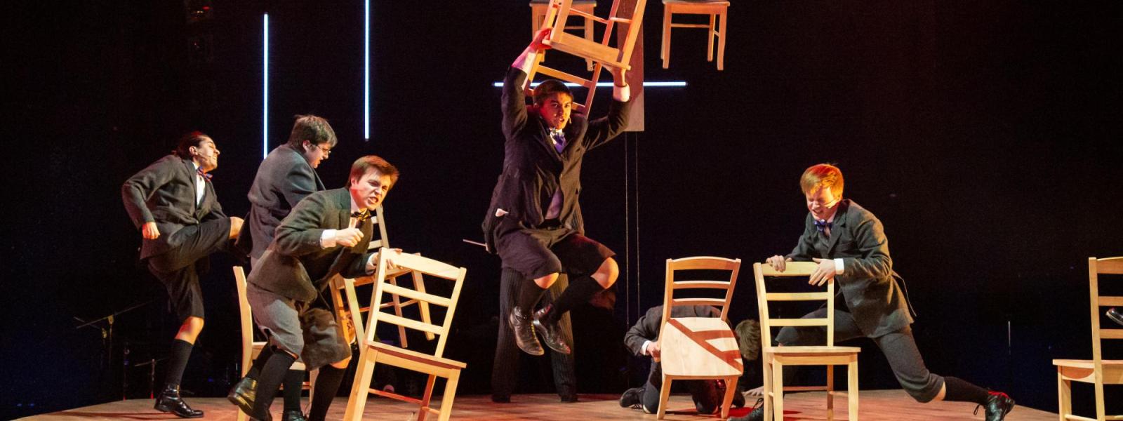 Student performers stamp around on stage and throw chairs