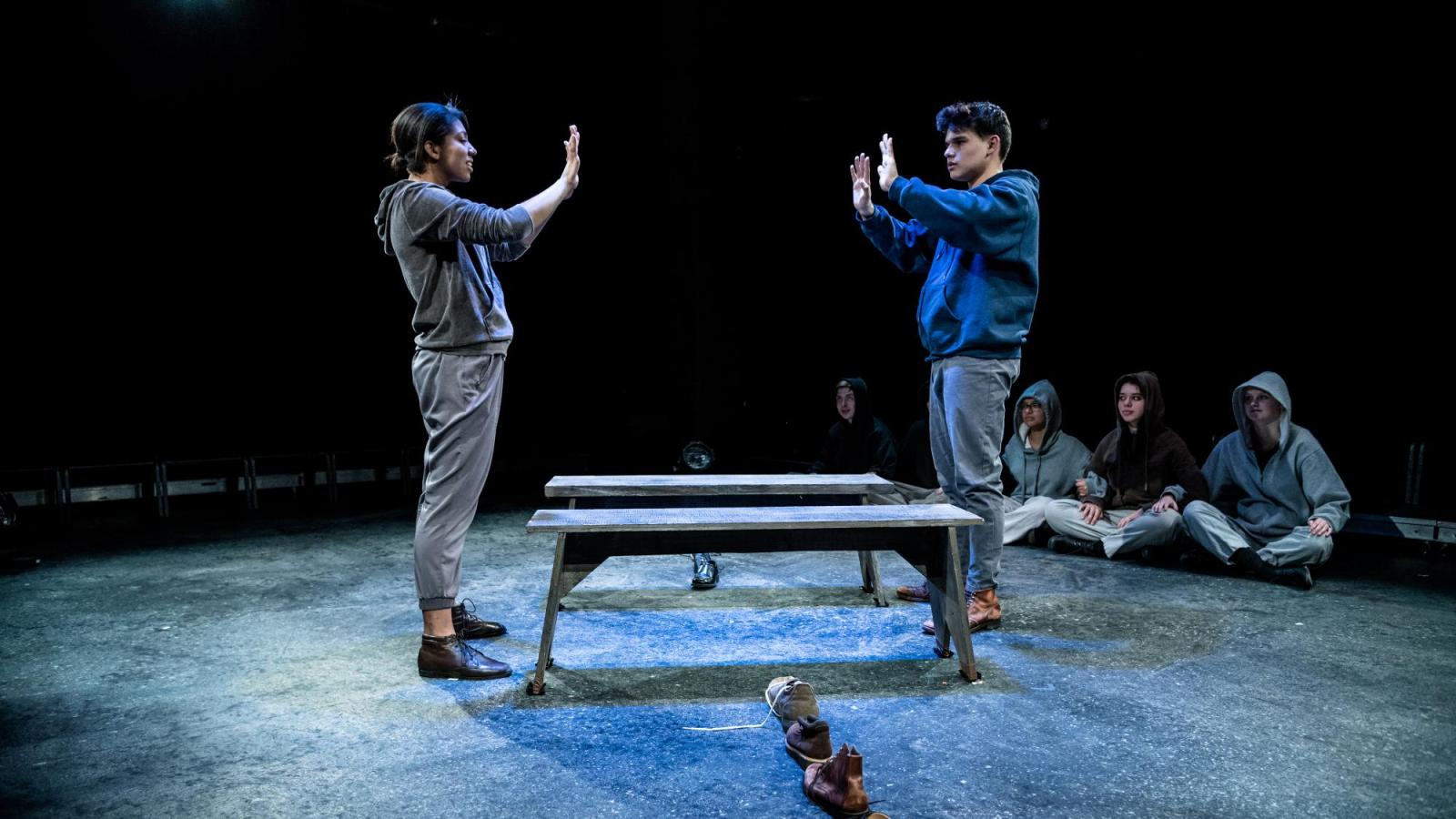 Two students stand apart with their hands up towards each other while a line of shoes in on the floor beneath them