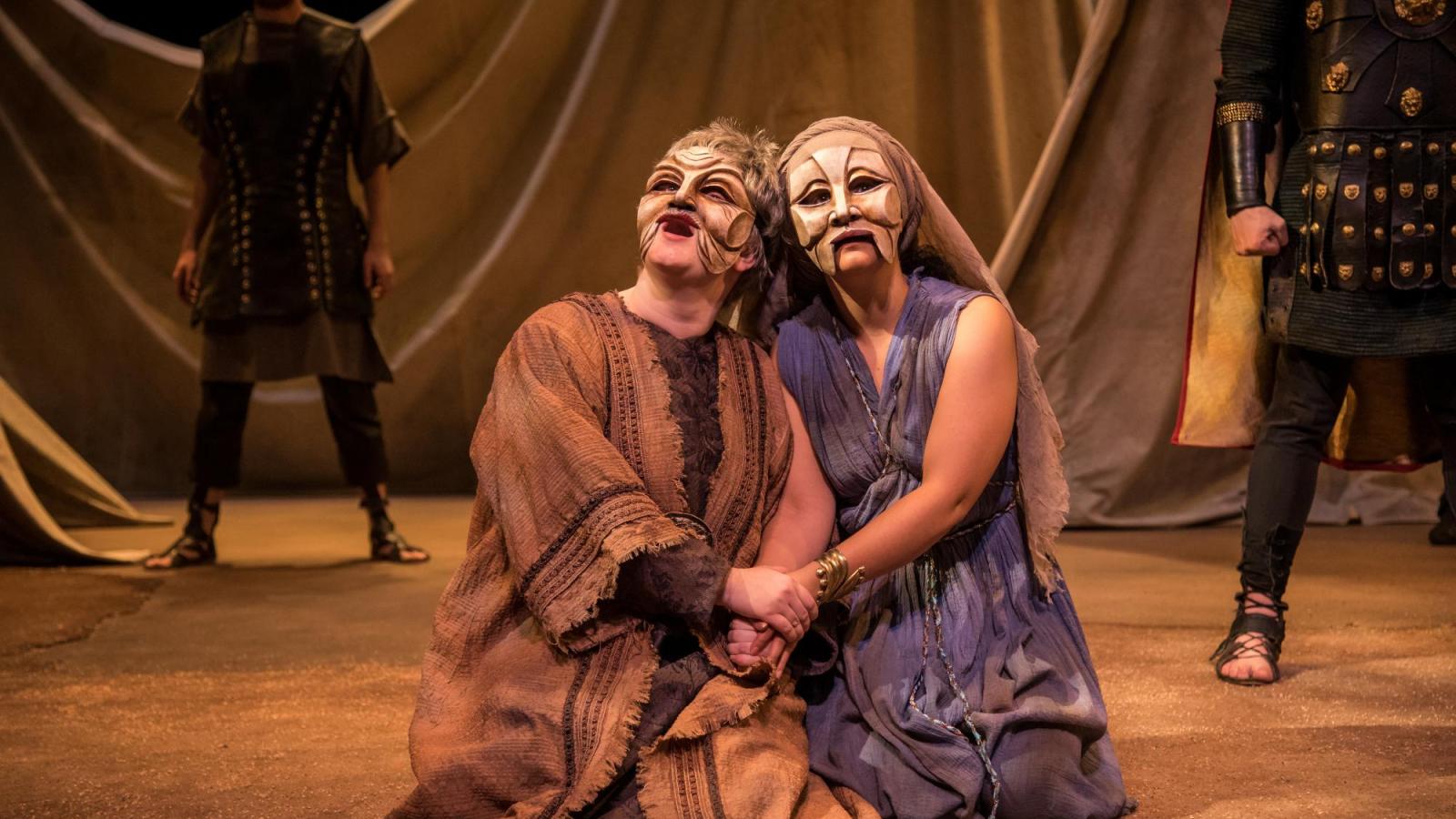 two masked students actors kneel and hold each other's hands