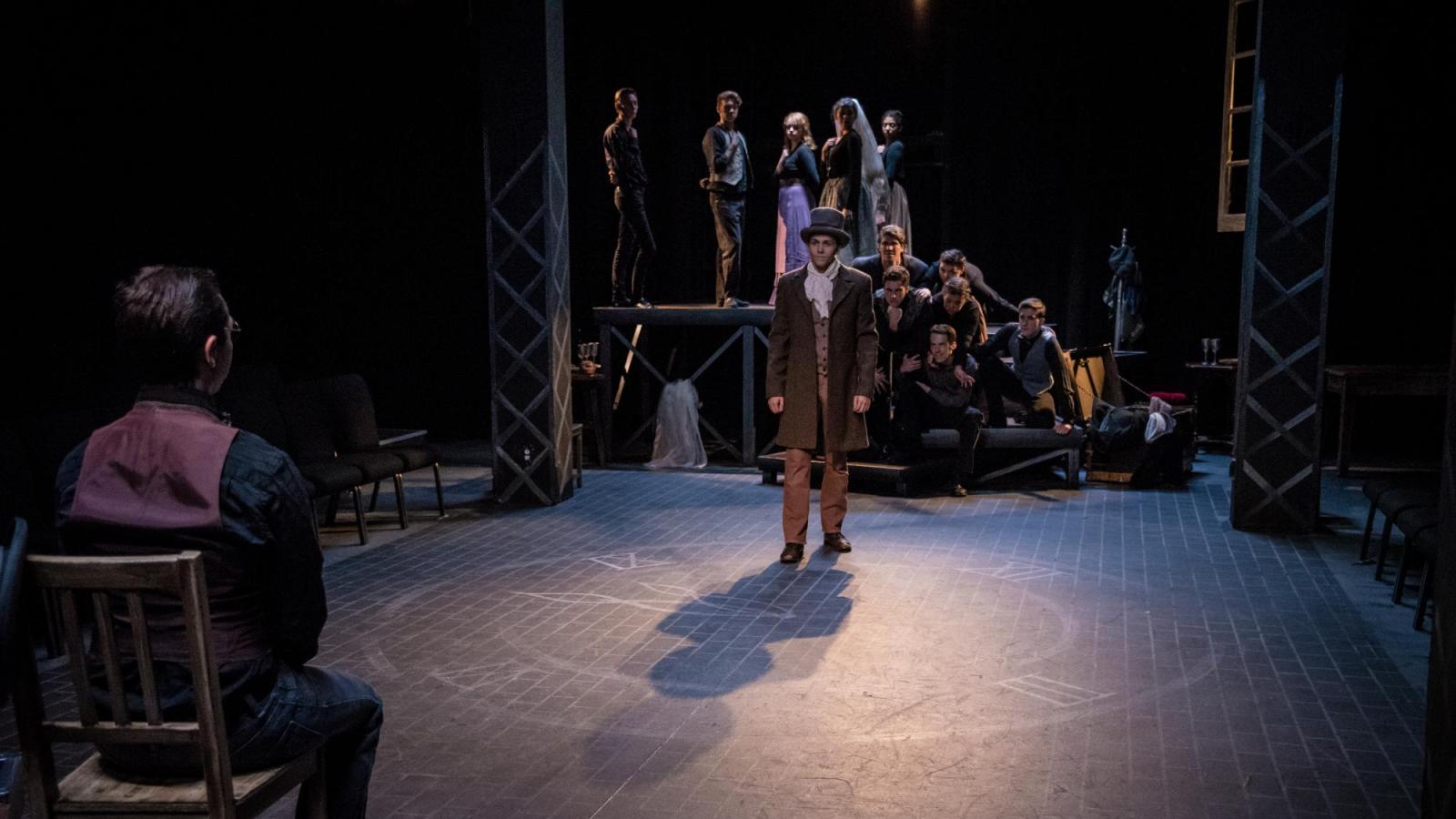 A student actor in a top hat approaches another who sits in a chair. Several others watch