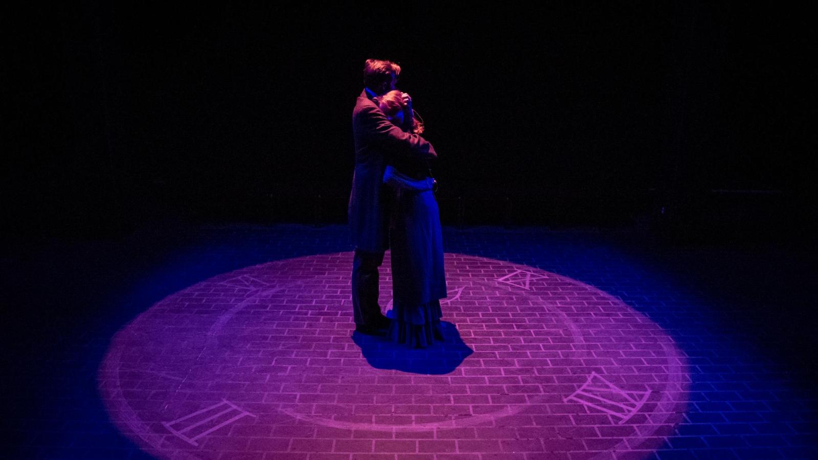 two actors embrace while standing on a clock face