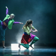 Dancers in bright purple, green, blue and red costumes under lighting