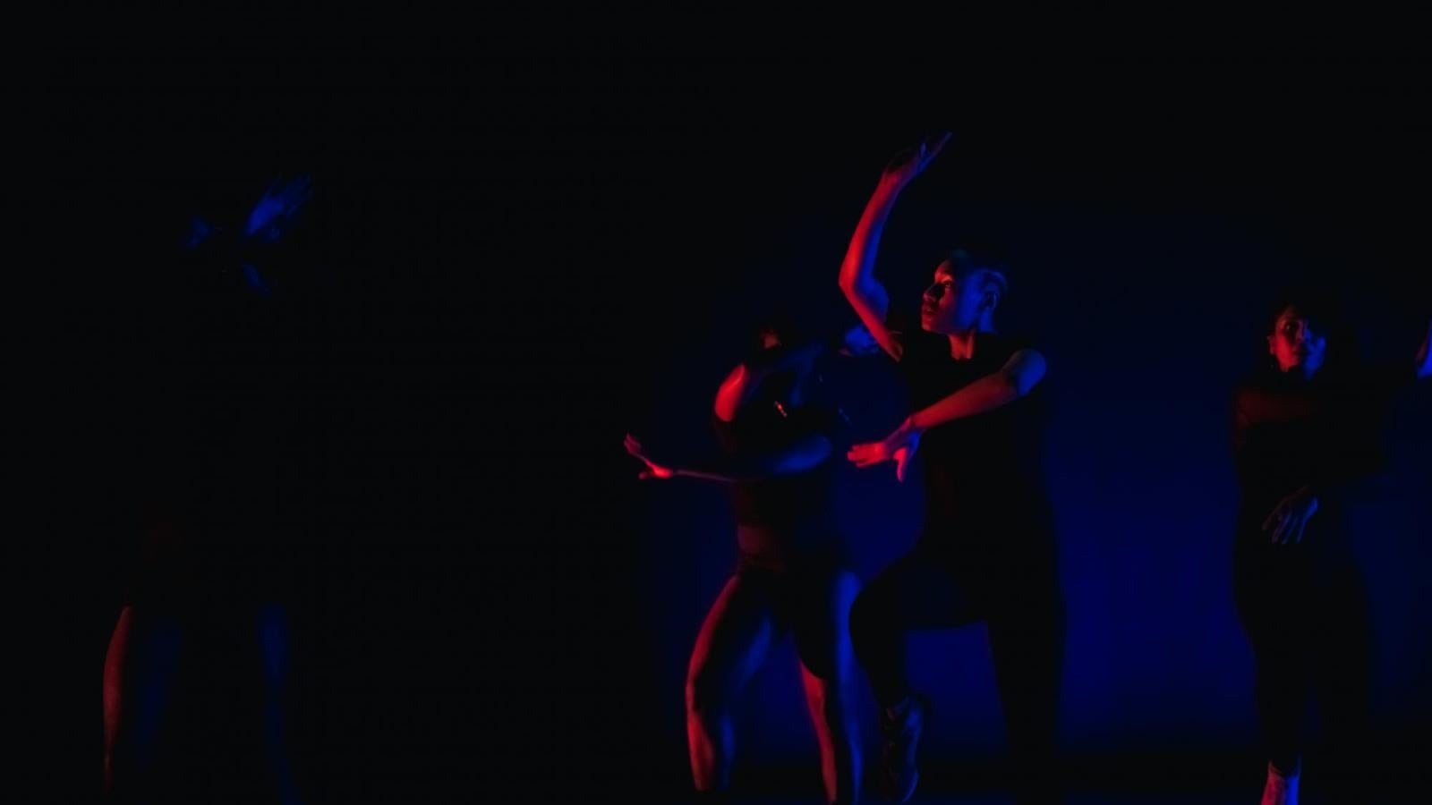 Several dancers perform on a darkly lit stage with red and blue lighting