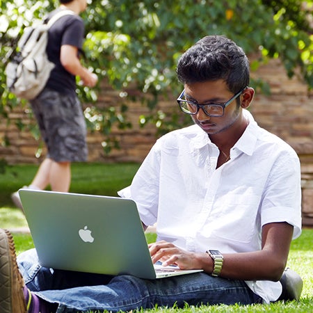 Student working on laptop, sitting in grass on campus