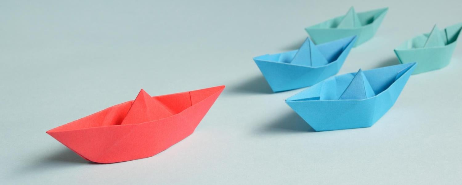 red paper boat leads a pack of blue and green paper boats