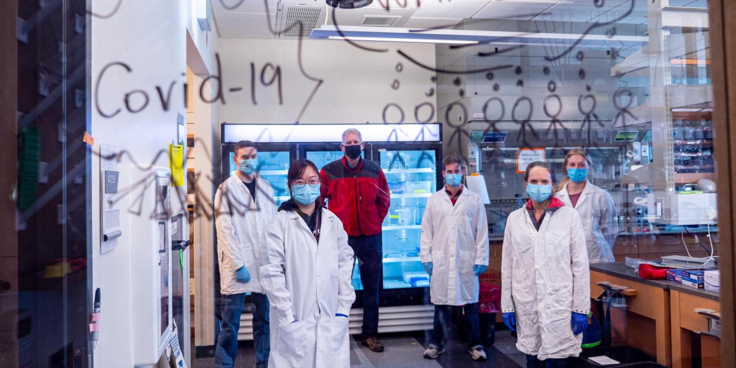 Researchers pose in their lab behind glass with the words "COVID Warriors" written on it.