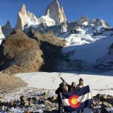 Three women hold a large Colorado flag and post for a photo on a mountain top in Patagonia.