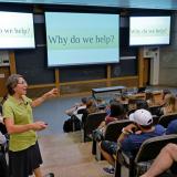 Wroe speaks to a group of students in a classroom, the screen says, "Why do we help?"