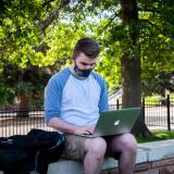 Student in mask works on laptop outside
