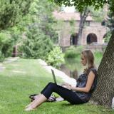 Grad student works on laptop while leaning against a tree