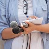 In a stock image, a journalist stands with two microphones, a reporter's notebook and pen.