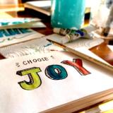 An illustration of the word "joy" is seen in a sketch book with a paintbrush nearby. (Photo by Bekka Mongeau from Pexels)