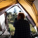 A man sitting in a tent, facing out, drinking from a cup