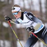 Nordic skier Hanna Abrahamsson wears a CU headband, sunglasses and a competition jersey, in a racing position with poles in hand, and small tree trunks in the background.