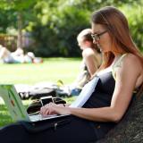 Student sits outside, leaning against tree, working on laptop