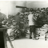 Black and white image of men looking through massive piles of books