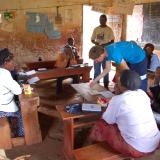 Man moves a piece on a wooden board, while several other people watch sitting at school desks