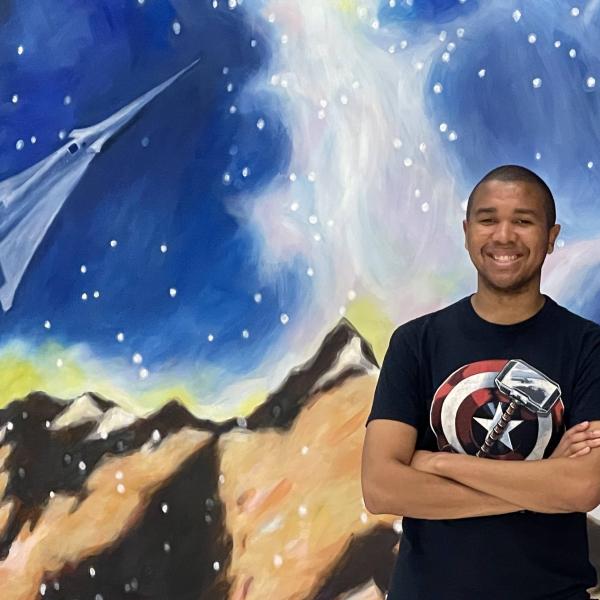 Dezell Turner stands with his arms folded in front of a mural showing mountains, a starry sky and a spaceshift flying above
