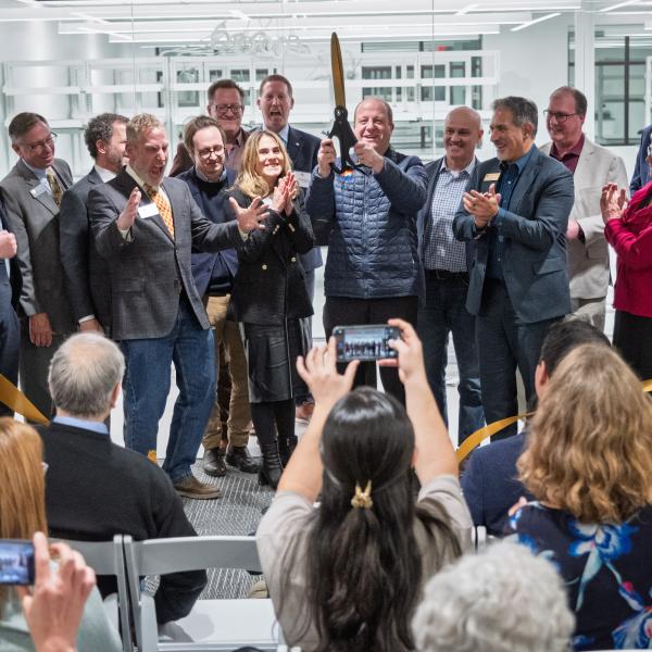 Platform guests and dignitaries watch Gov. Jared Polis celebrate after cutting a ribbon to signify the opening of the Quantum Incubator site