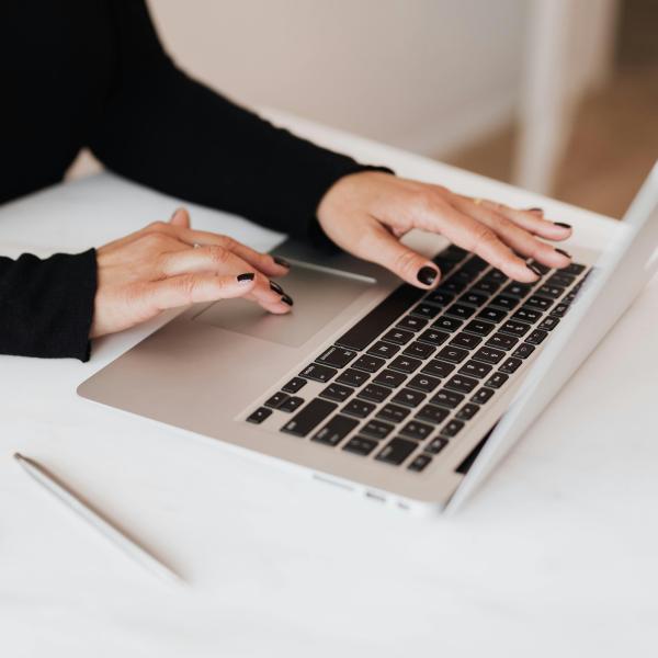 Arms in a black sweater extend to a laptop keyboard, with a phone and pen on the table. (Photo: Pexels)