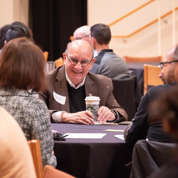 Phililp DiStefano sits at a table with others, smiling, to-go cup in hand.