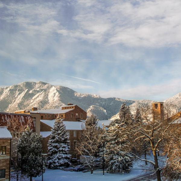 Campus buildings and the Flatirons in the background are frosted with snow, under a partly sunny sky—colors of blue brown and white.