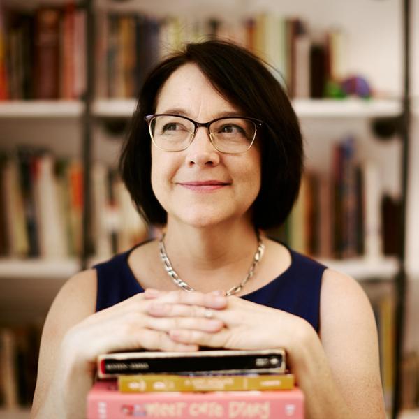 Woman crosses hands over a pile of books with a bookshelf in the backgroundbra