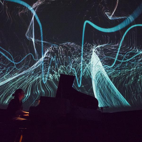 A person is on a dark stage with multi-colored laser light designs as the backdrop.