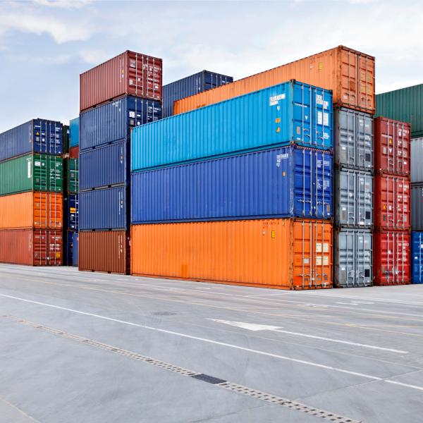 More than a dozen shipping crates stacked on dry ground, likely in a port. The crates are blue, orange, green and gray.