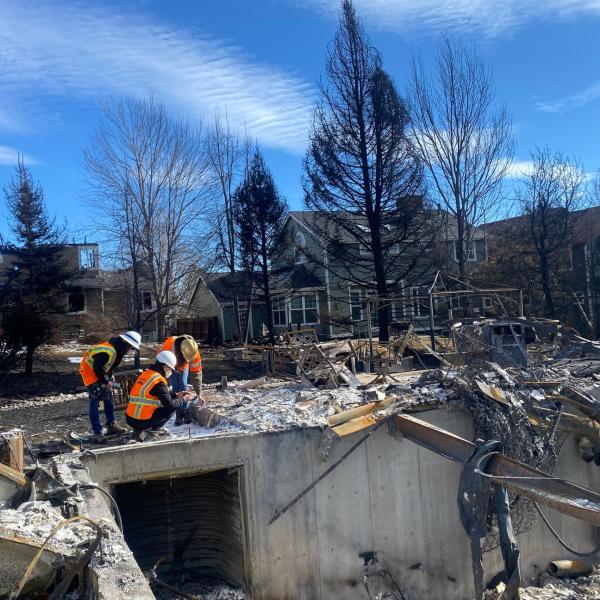 Three people in neon vests kneeling amid rubble from the Marshall Fire