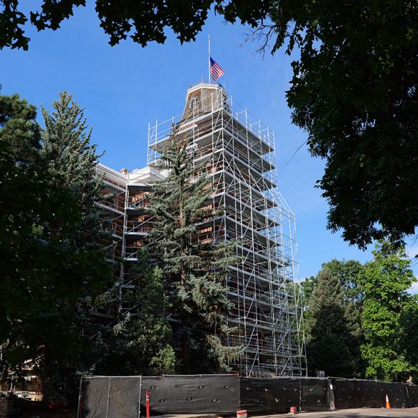 Framed by tree leaves and evergreens, a view of the Old Main tower surrounded by scaffolding and topped with the American flag.